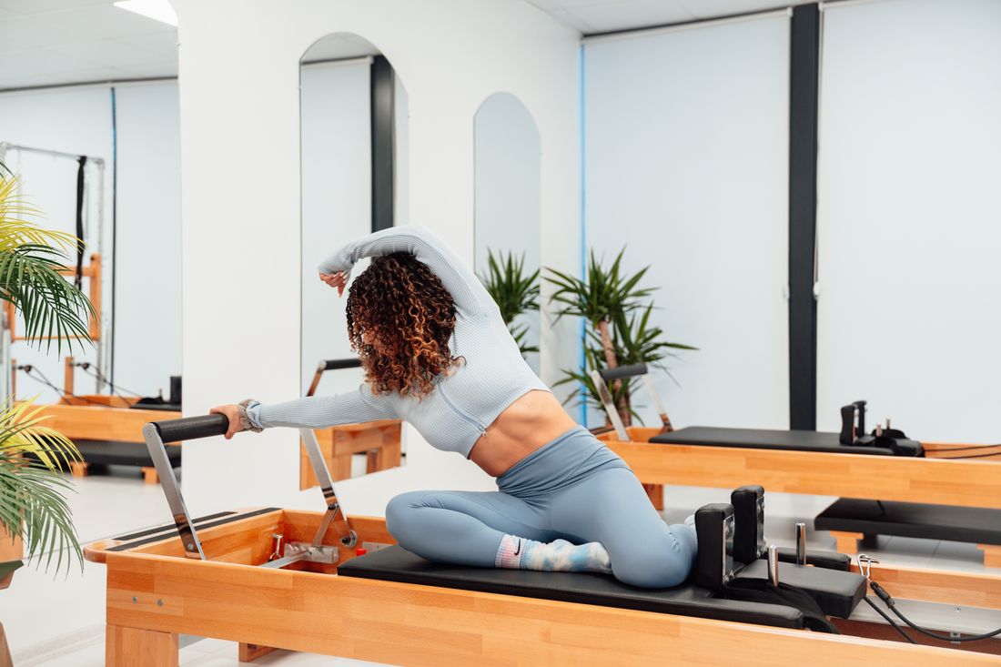 Mulher praticando pilates no reformer, alongando-se com blusa cinza sobre o aparelho